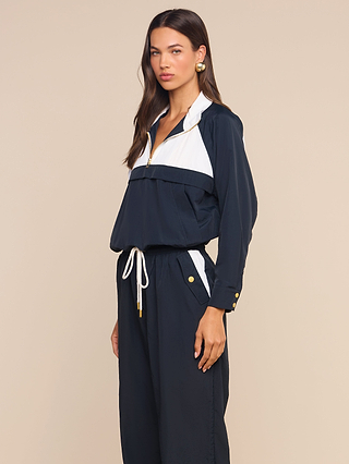 A woman stands against a beige background, wearing a navy blue and white long-sleeve jumpsuit with gold buttons and a drawstring waist, styled beneath the Vex Cropped Windbreaker for a modern layered look, looking at the camera.