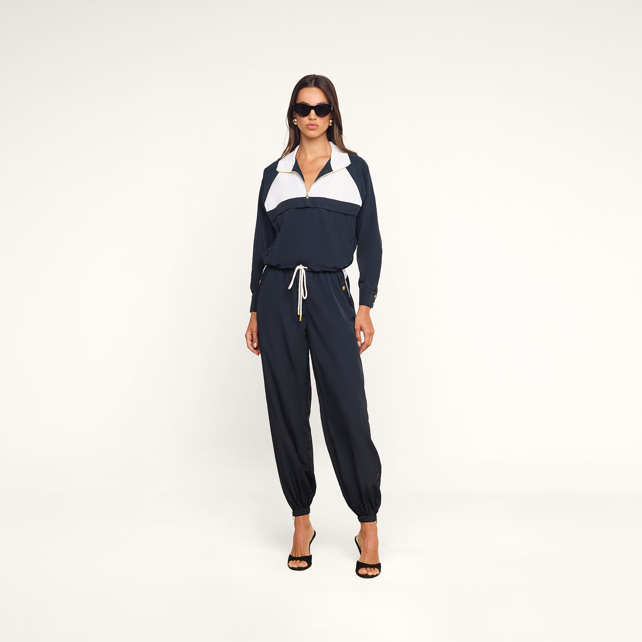 A woman stands confidently against a plain white background, hands in her pockets and wearing sunglasses, styled in the Vex Cropped Windbreaker and black heels.