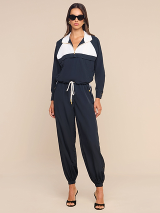 A woman stands against a beige background wearing the Vex Cropped Windbreaker—a navy blue and white water-repellent activewear jacket with a drawstring waist—paired with black high-heeled sandals, sunglasses, and gold earrings.