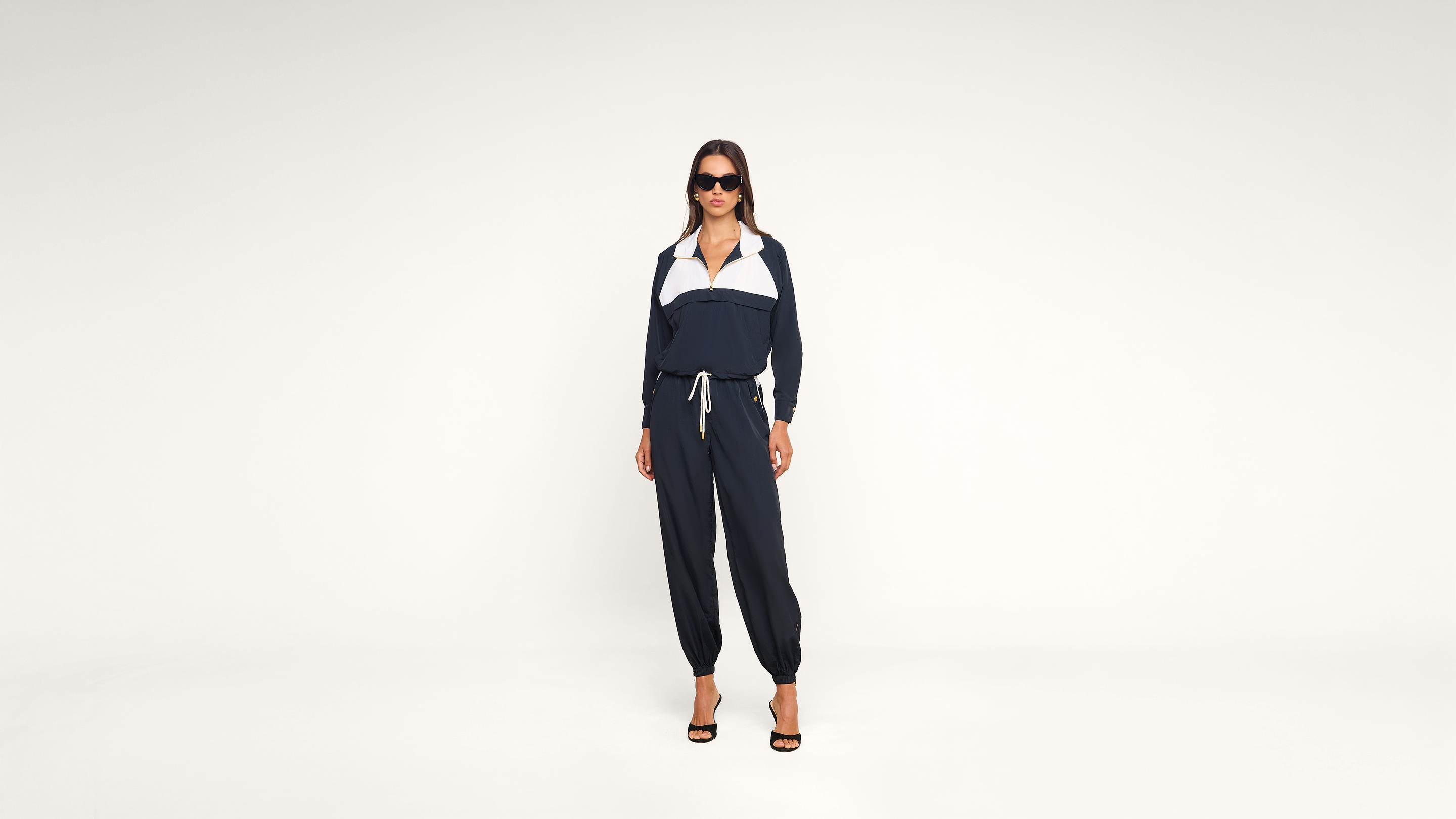 A woman wearing sunglasses and a navy blue tracksuit with white accents stands confidently in heels against a plain white background.