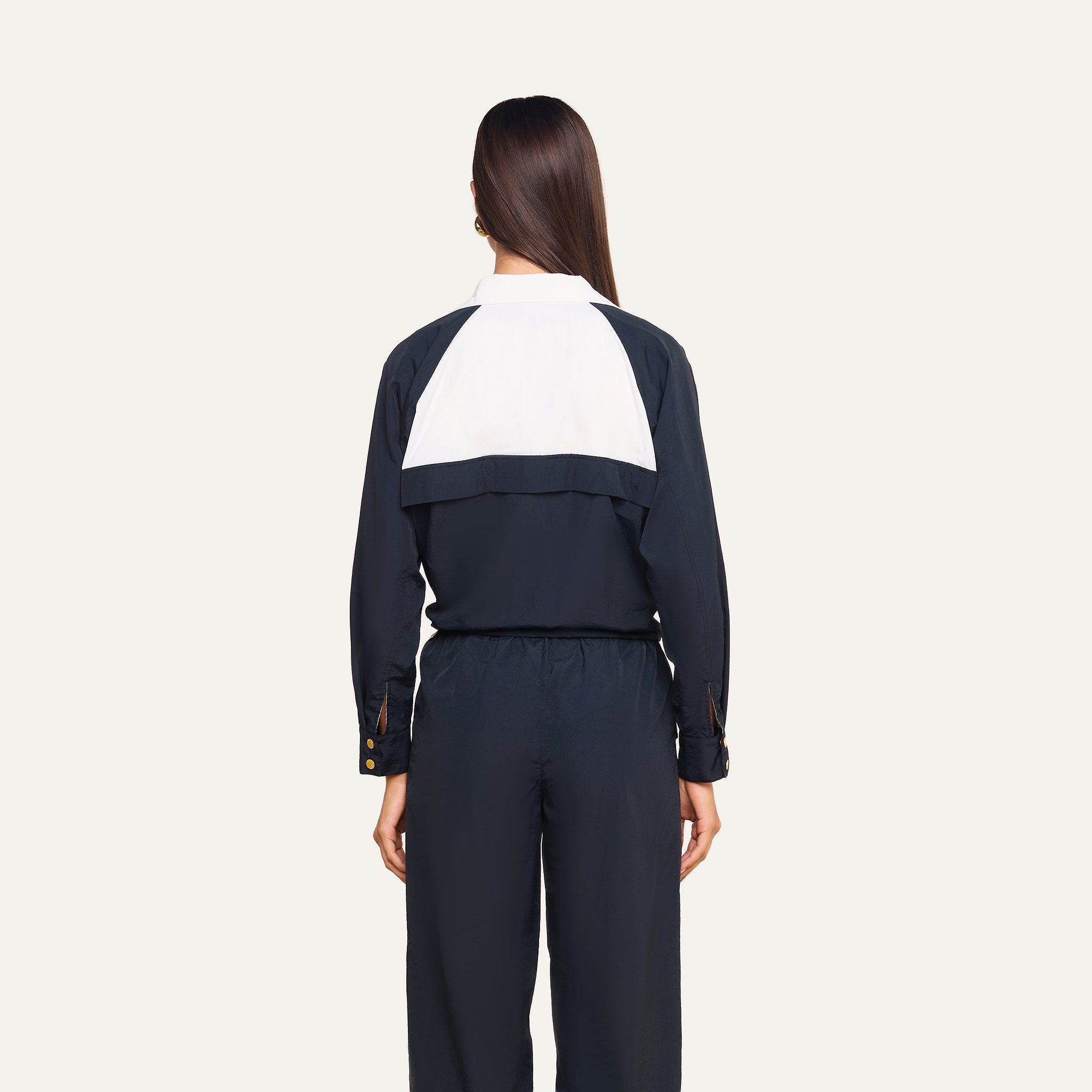 A woman with straight brown hair, wearing a black jumpsuit featuring a white panel on the back, stands facing away against a plain light background, paired with the Vex Cropped Windbreaker.