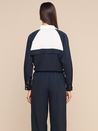 A person with long brown hair, seen from behind, wears a navy blue jumpsuit with a white panel on the upper back and brown button details on the sleeves, layered under the Vex Cropped Windbreaker—an activewear jacket designed to be both water-repellent and stylish.