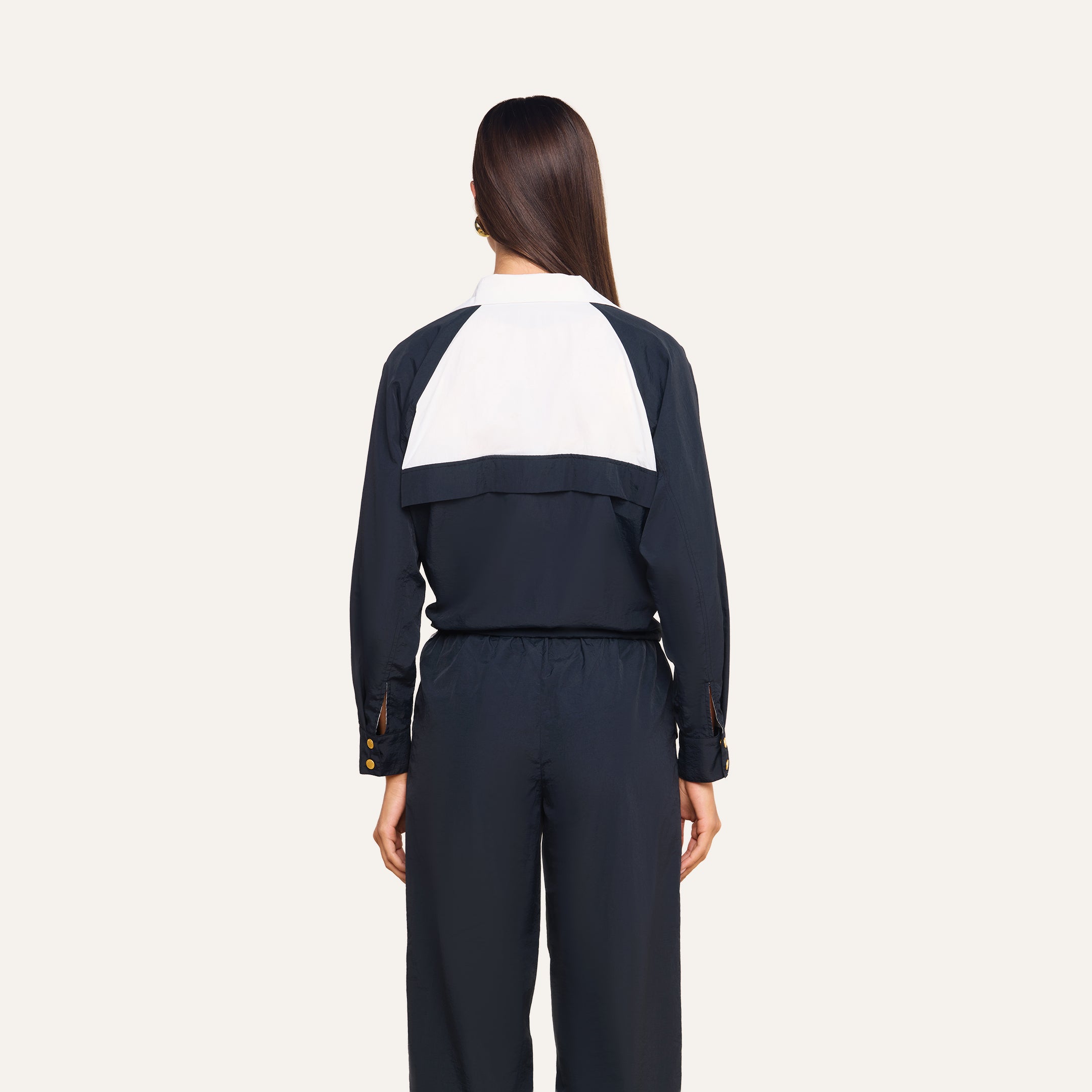 A woman with straight brown hair, wearing a black jumpsuit featuring a white panel on the back, stands facing away against a plain light background, paired with the Vex Cropped Windbreaker.