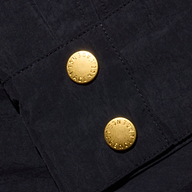 Close-up of the Vex Cropped Windbreaker’s dark, water-repellent fabric with two gold buttons, each engraved with the word “AGENCY” in a circular pattern around the edge.