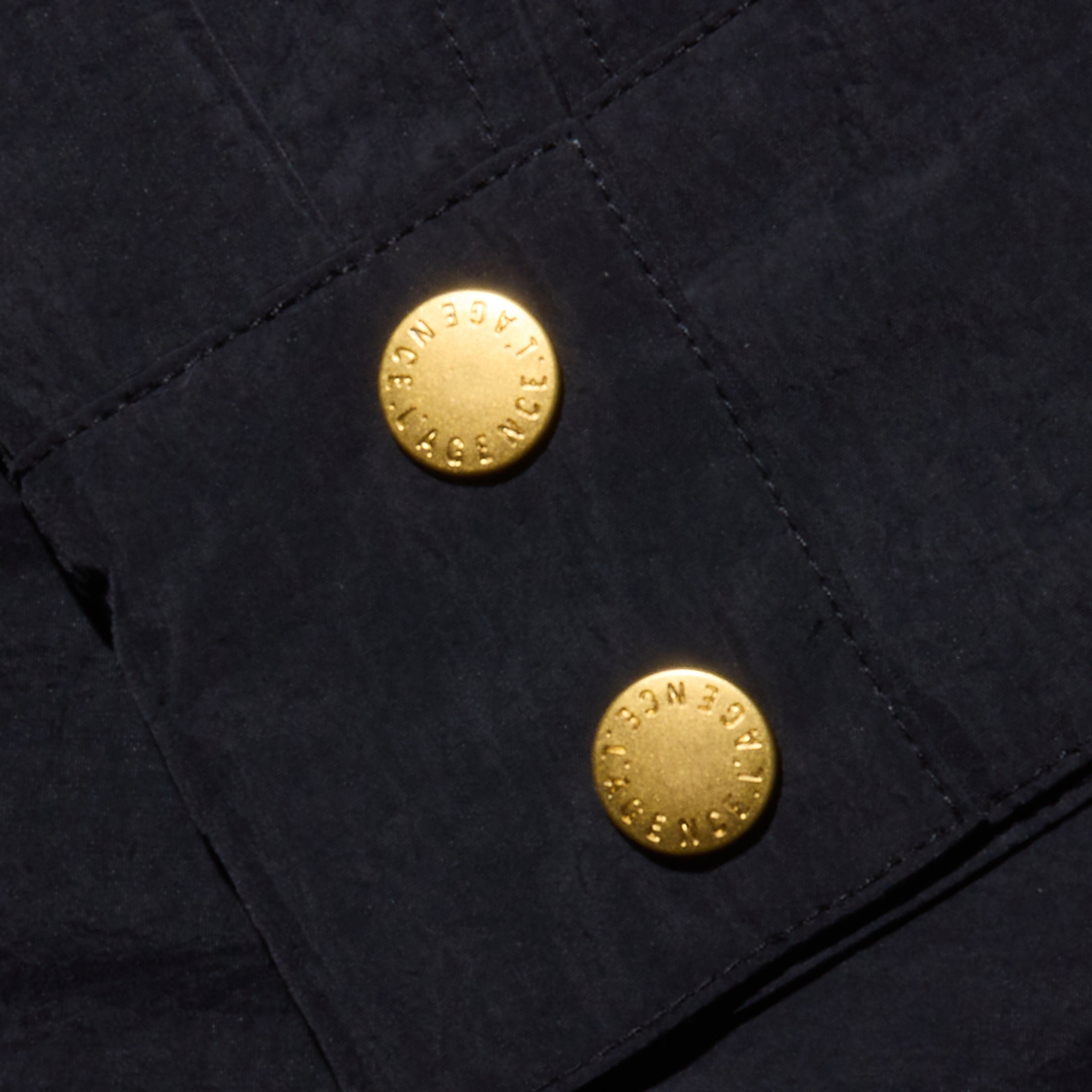 Close-up of the Vex Cropped Windbreaker’s dark, water-repellent fabric with two gold buttons, each engraved with the word “AGENCY” in a circular pattern around the edge.