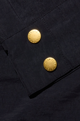 Close-up of the Vex Cropped Windbreaker’s dark fabric sleeve, featuring two shiny gold buttons embossed with fine lettering and fastened to a rectangular cuff. The material has a textured look, characteristic of durable cotton or denim—emphasizing the garment’s activewear-inspired windbreaker design.