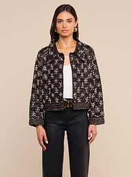 Una donna che indossa la giacca in pelle decorata Morrisa — una giacca nera decorata a mano con un motivo geometrico bianco — abbinata a un top bianco, pantaloni neri e orecchini a cerchio, è in posa su uno sfondo beige.