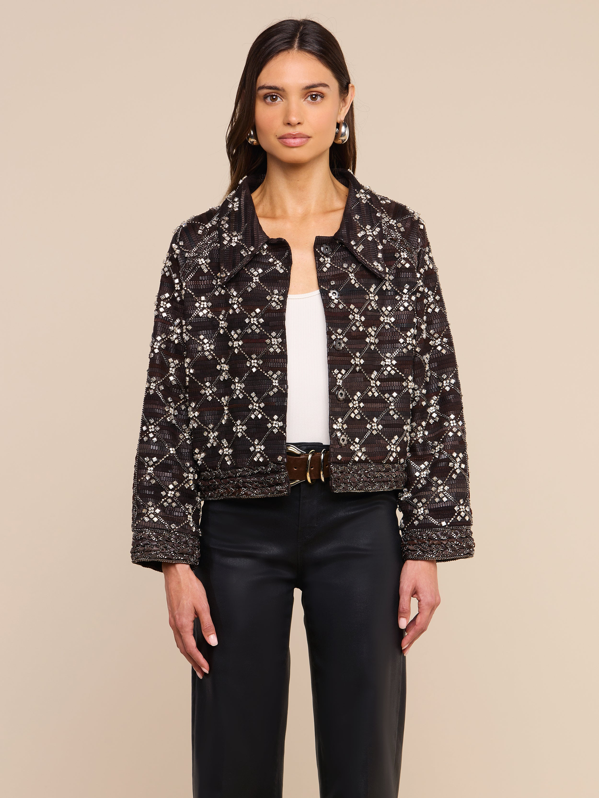 Una donna che indossa la giacca in pelle decorata Morrisa — una giacca nera decorata a mano con un motivo geometrico bianco — abbinata a un top bianco, pantaloni neri e orecchini a cerchio, è in posa su uno sfondo beige.