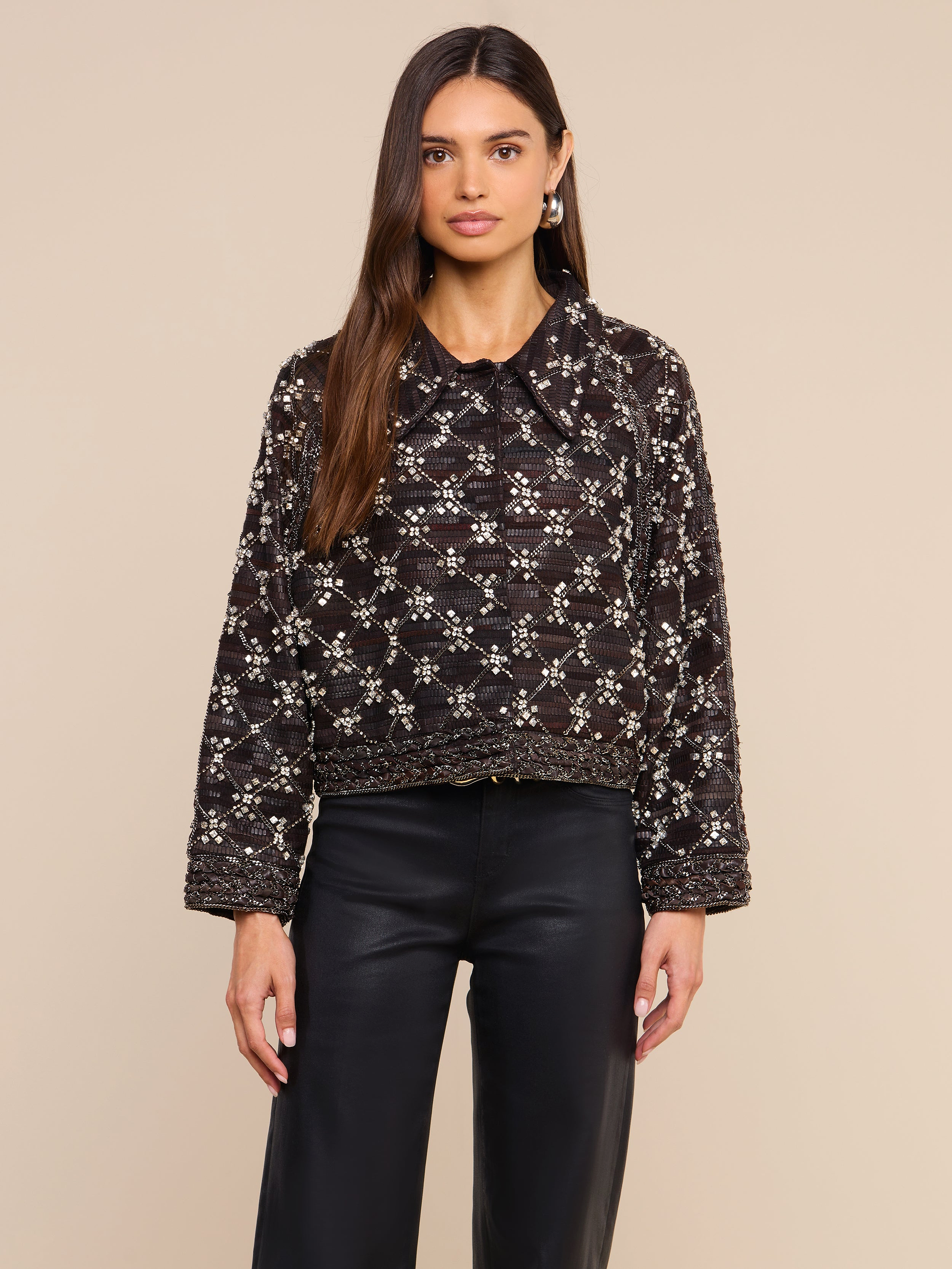 Una donna dai lunghi capelli castani indossa la giacca in pelle decorata Morrisa — caratterizzata da un suggestivo motivo geometrico bianco realizzato con perline — e pantaloni in pelle neri, in piedi su uno sfondo beige.
