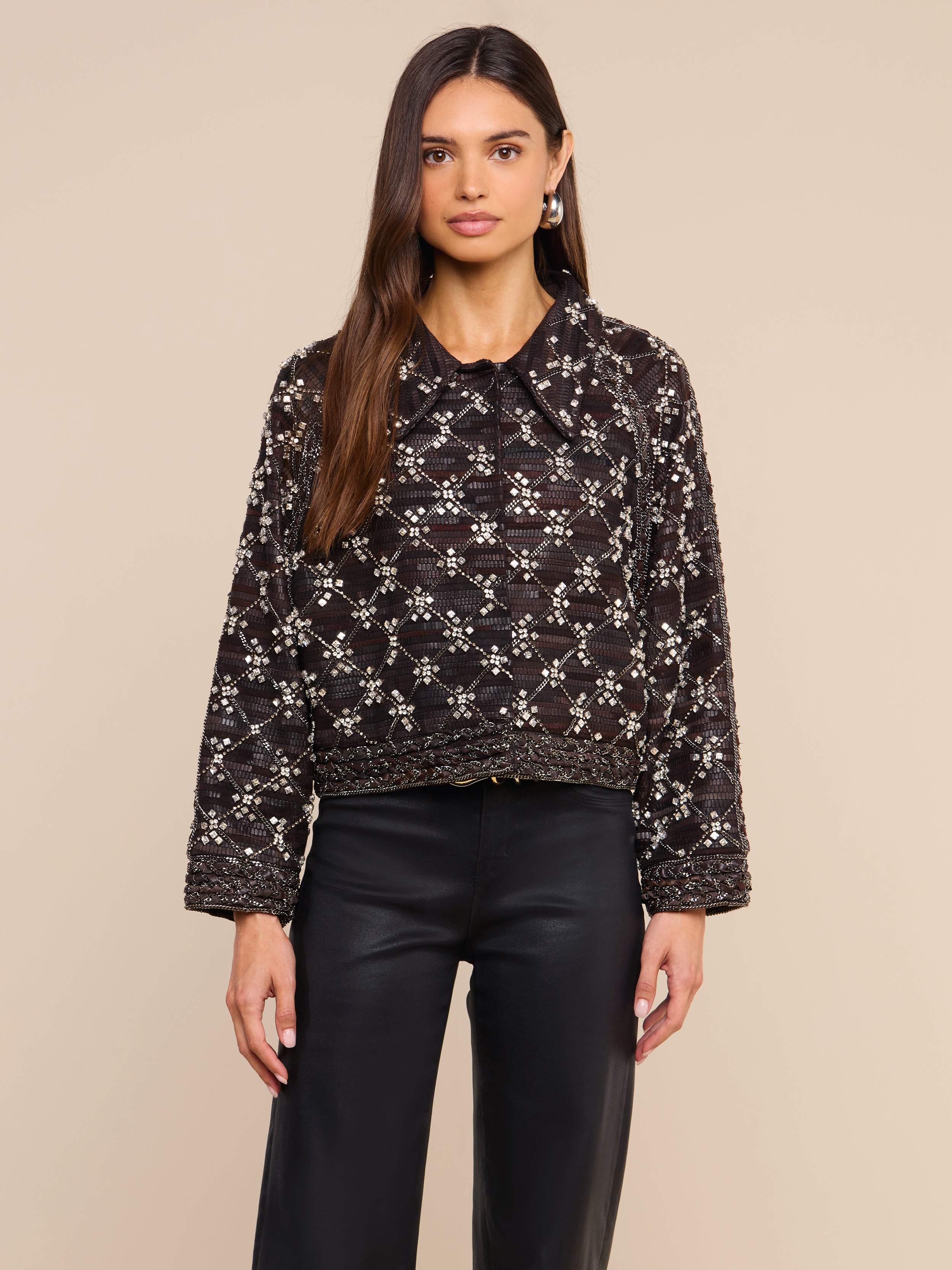 Una donna dai lunghi capelli castani indossa la giacca in pelle decorata Morrisa — caratterizzata da un suggestivo motivo geometrico bianco realizzato con perline — e pantaloni in pelle neri, in piedi su uno sfondo beige.