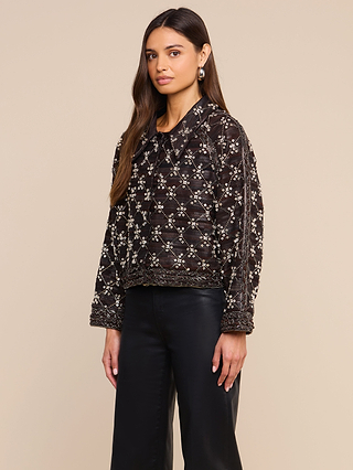 A woman with long brown hair wears the Morrisa Embellished Leather Jacket, crafted from recycled leather, paired with black pants. She stands against a beige background, looking toward the camera with a neutral expression—a reflection of her commitment to sustainable fashion.