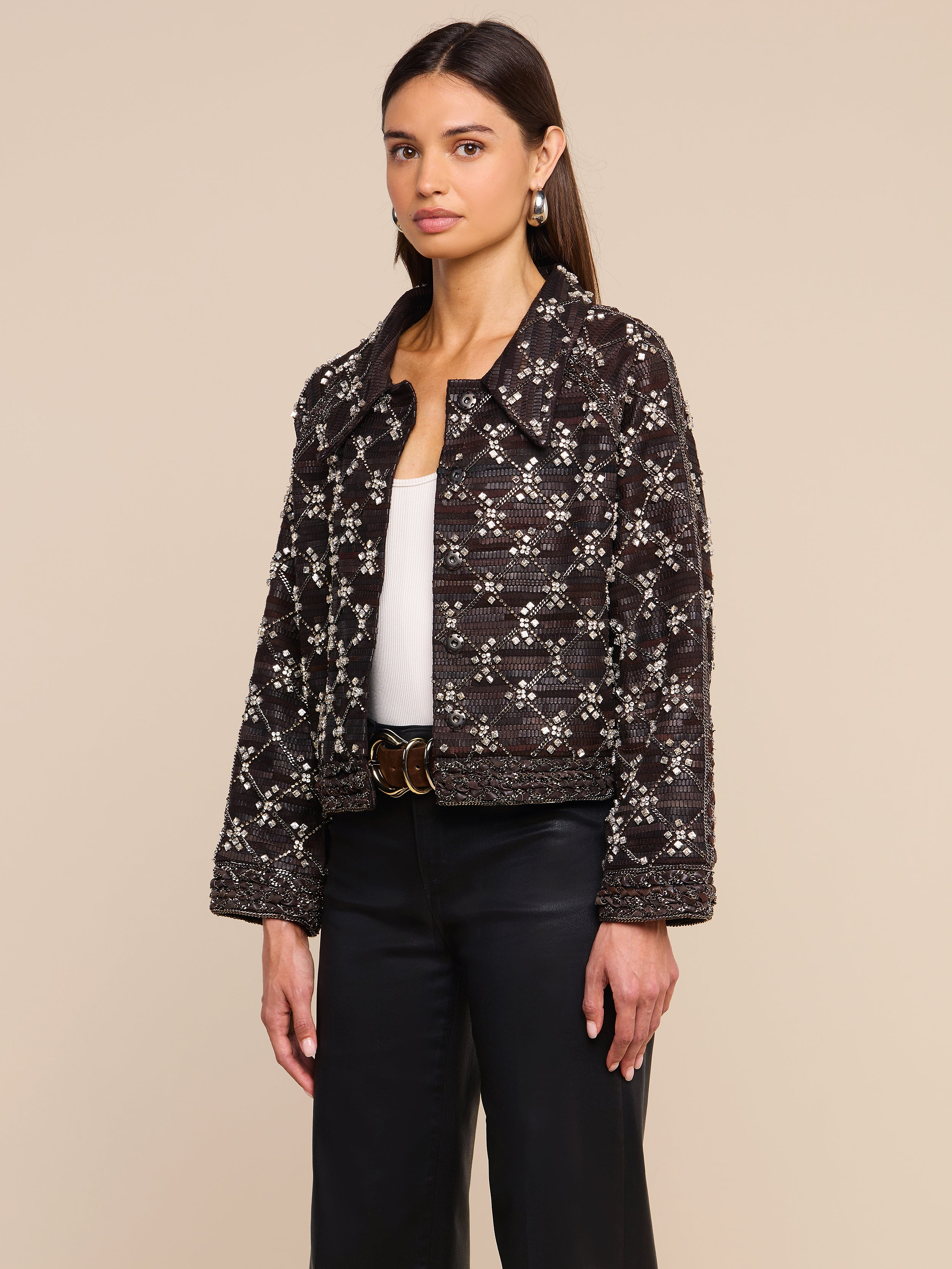 Una donna che indossa la giacca in pelle decorata Morrisa, realizzata in pelle nera con intricati motivi bianchi, abbinata a un top bianco e pantaloni neri, è ritratta su uno sfondo beige a tinta unita.