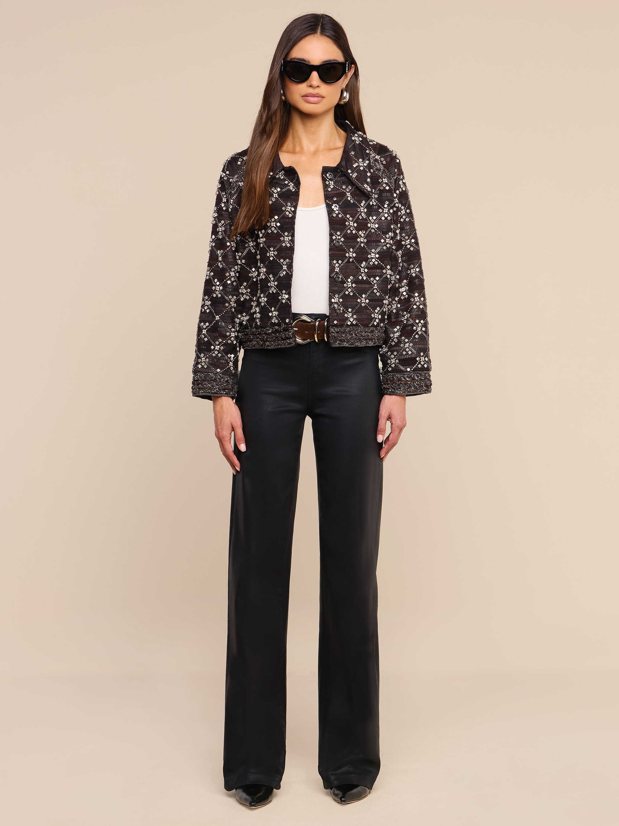 Una donna con indosso occhiali da sole, la giacca in pelle decorata Morrisa con motivo nero, un top bianco, pantaloni neri a gamba larga e scarpe nere è ritratta di fronte, su uno sfondo beige a tinta unita.