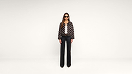 A woman stands against a plain white background, wearing black sunglasses, a patterned jacket, a white top, black wide-leg pants, and black shoes.