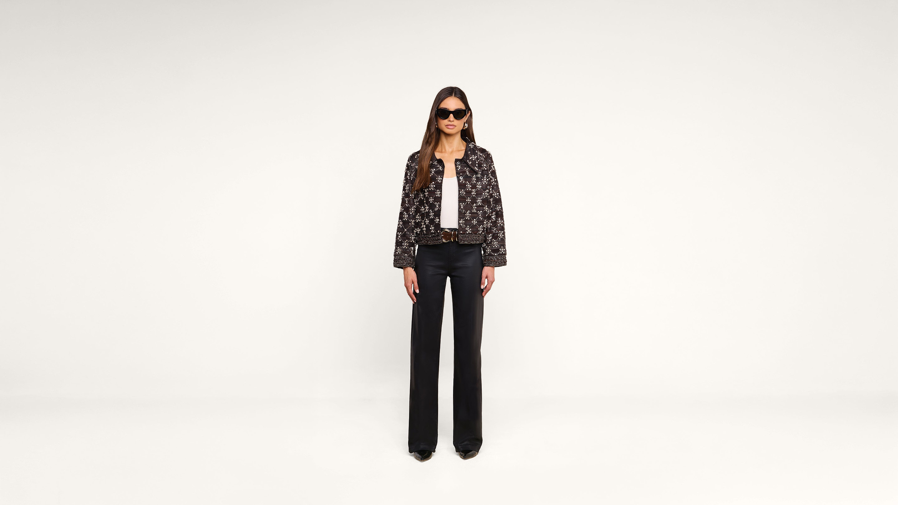 A woman stands against a plain white background, wearing black sunglasses, a patterned jacket, a white top, black wide-leg pants, and black shoes.
