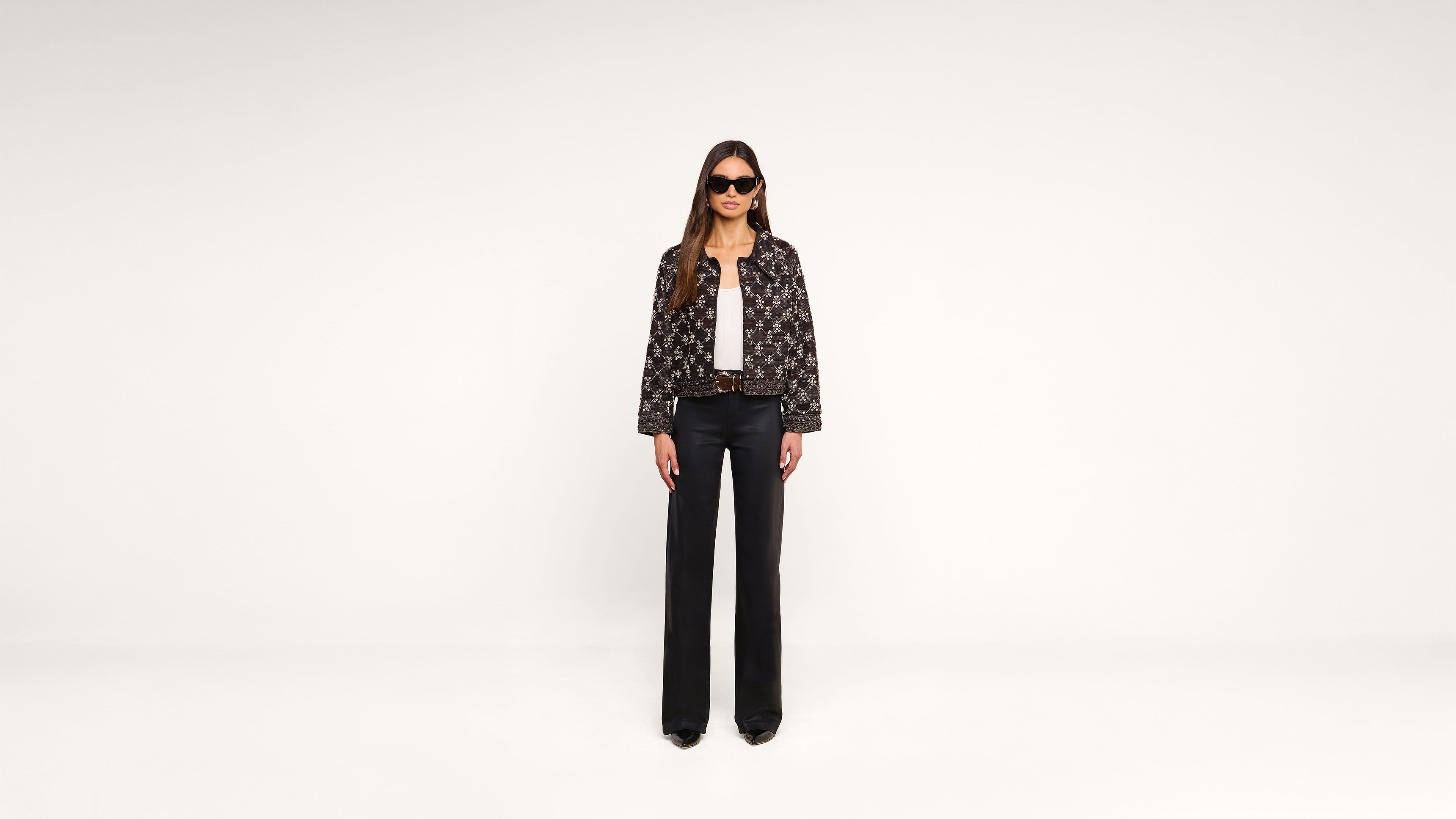 A woman stands against a plain white background, wearing black sunglasses, a patterned jacket, a white top, black wide-leg pants, and black shoes.