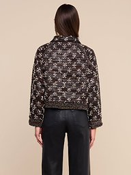 Una persona con lunghi capelli castani, vista di spalle, indossa la giacca in pelle decorata Morrisa, con motivi nei toni del marrone scuro e del bianco, abbinata a pantaloni in pelle neri, in piedi su uno sfondo beige. Questo capo di grande impatto mette in risalto la maestria artistica della moda sostenibile.