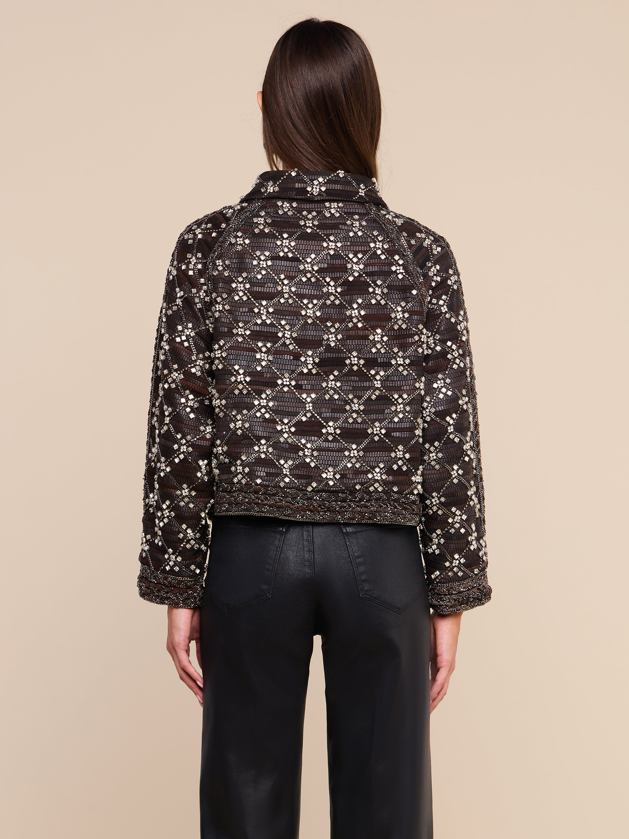 Una persona con lunghi capelli castani, vista di spalle, indossa la giacca in pelle decorata Morrisa, con motivi nei toni del marrone scuro e del bianco, abbinata a pantaloni in pelle neri, in piedi su uno sfondo beige. Questo capo di grande impatto mette in risalto la maestria artistica della moda sostenibile.