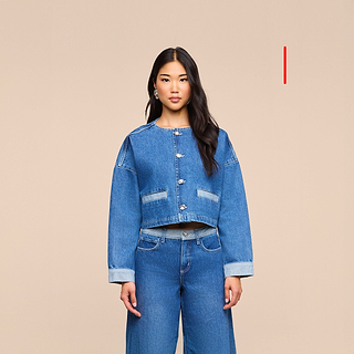 A woman with long dark hair wears the blue denim L’AGENCE Arabell Romper, standing against a plain beige background.