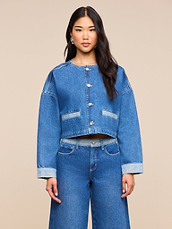 Una donna dai lunghi capelli scuri indossa la giacca di jeans Verona dal taglio oversize e i jeans a gamba larga abbinati, in piedi davanti a uno sfondo beige a tinta unita.