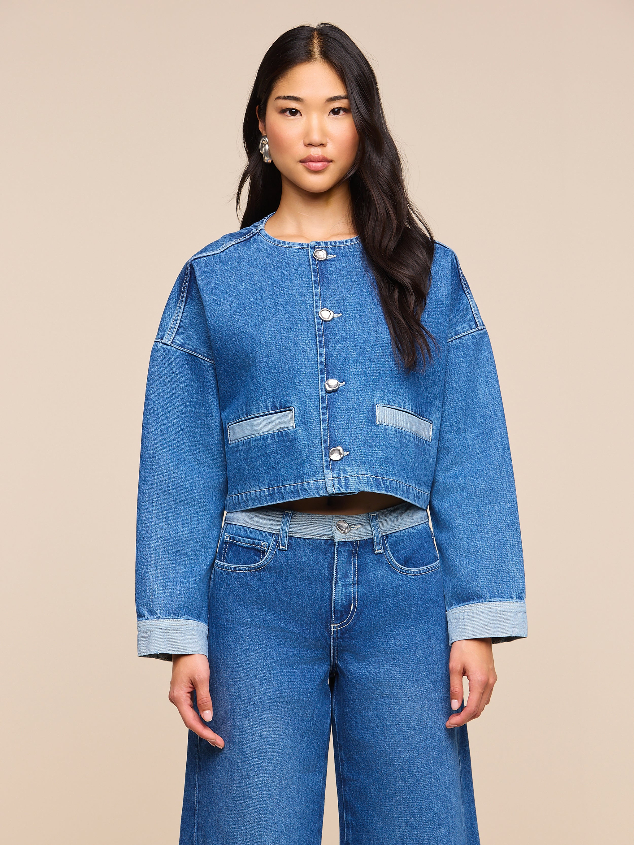 Una donna dai lunghi capelli scuri indossa la giacca di jeans Verona dal taglio oversize e i jeans a gamba larga abbinati, in piedi davanti a uno sfondo beige a tinta unita.