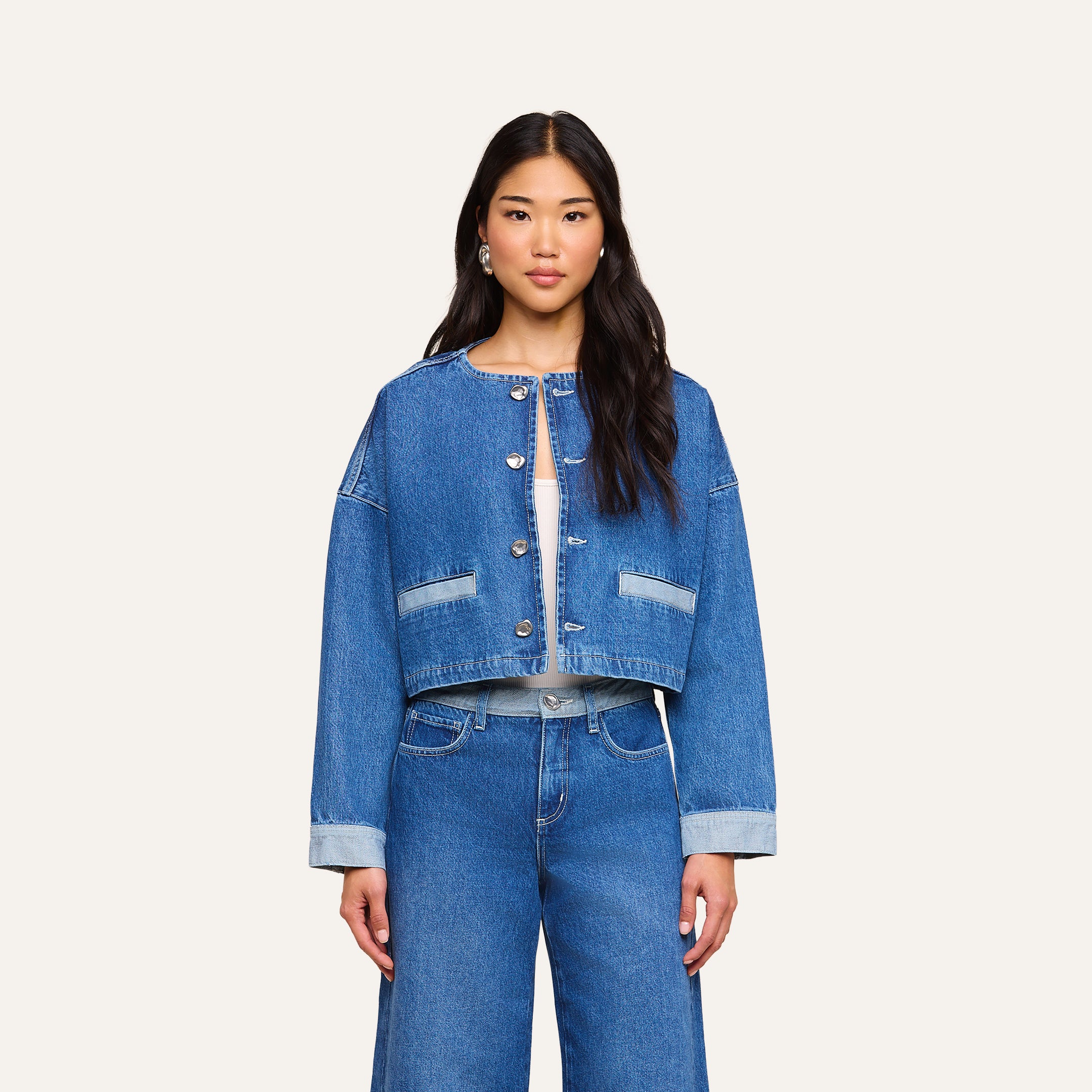 Una donna dai lunghi capelli scuri e dallespressione neutra è in piedi, rivolta in avanti, e indossa la giacca Verona Jean Jacket — una giacca di jeans blu senza colletto con bottoni argentati — abbinata a jeans a vita alta dello stesso modello. Lo sfondo è di un beige uniforme.