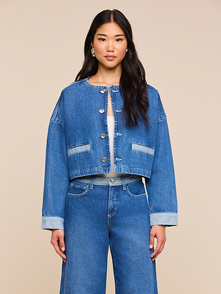 A woman with long dark hair and a neutral expression stands facing forward, wearing the Verona Jean Jacket— a blue, collarless denim jacket featuring silver buttons—paired with matching high-waisted jeans. The background is plain beige.