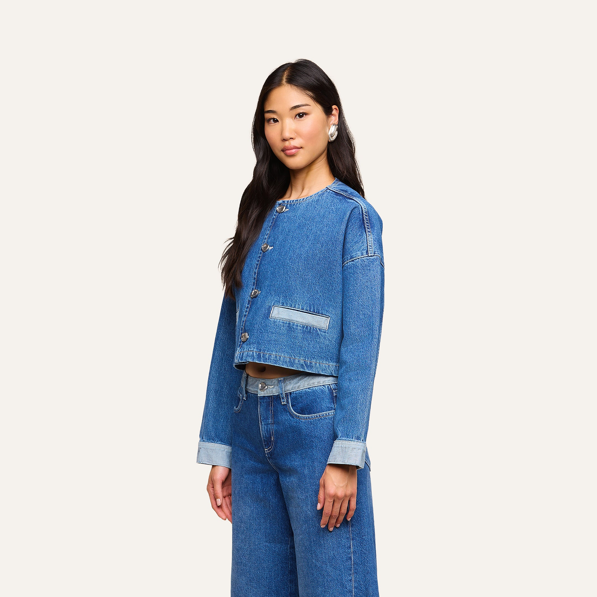 Una donna dai lunghi capelli scuri indossa la giacca Verona Jean Jacket e i jeans a vita alta abbinati, in piedi davanti a uno sfondo beige a tinta unita.