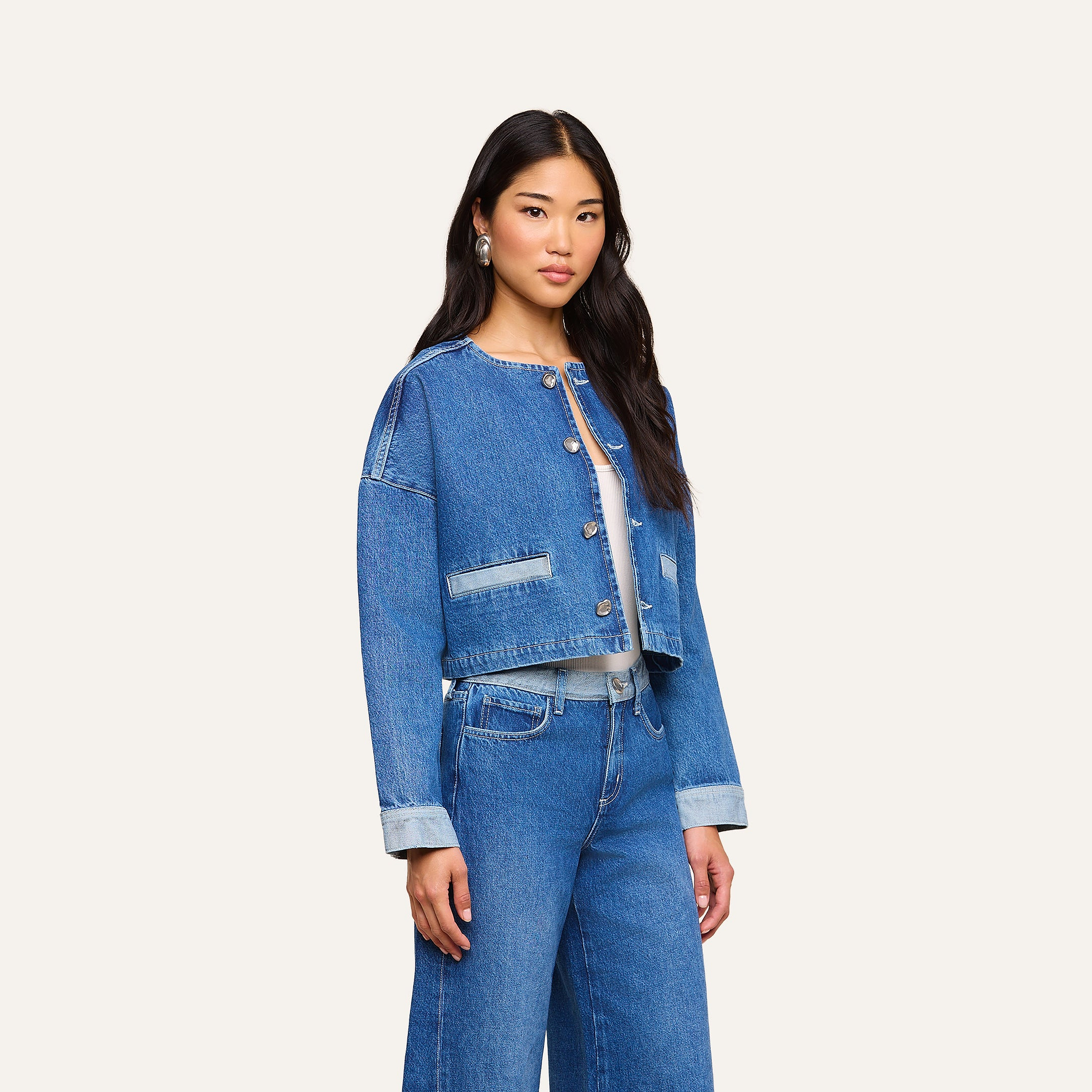 Una donna dai lunghi capelli scuri indossa la giacca Verona Jean e dei jeans blu a vita alta, in piedi davanti a uno sfondo beige.