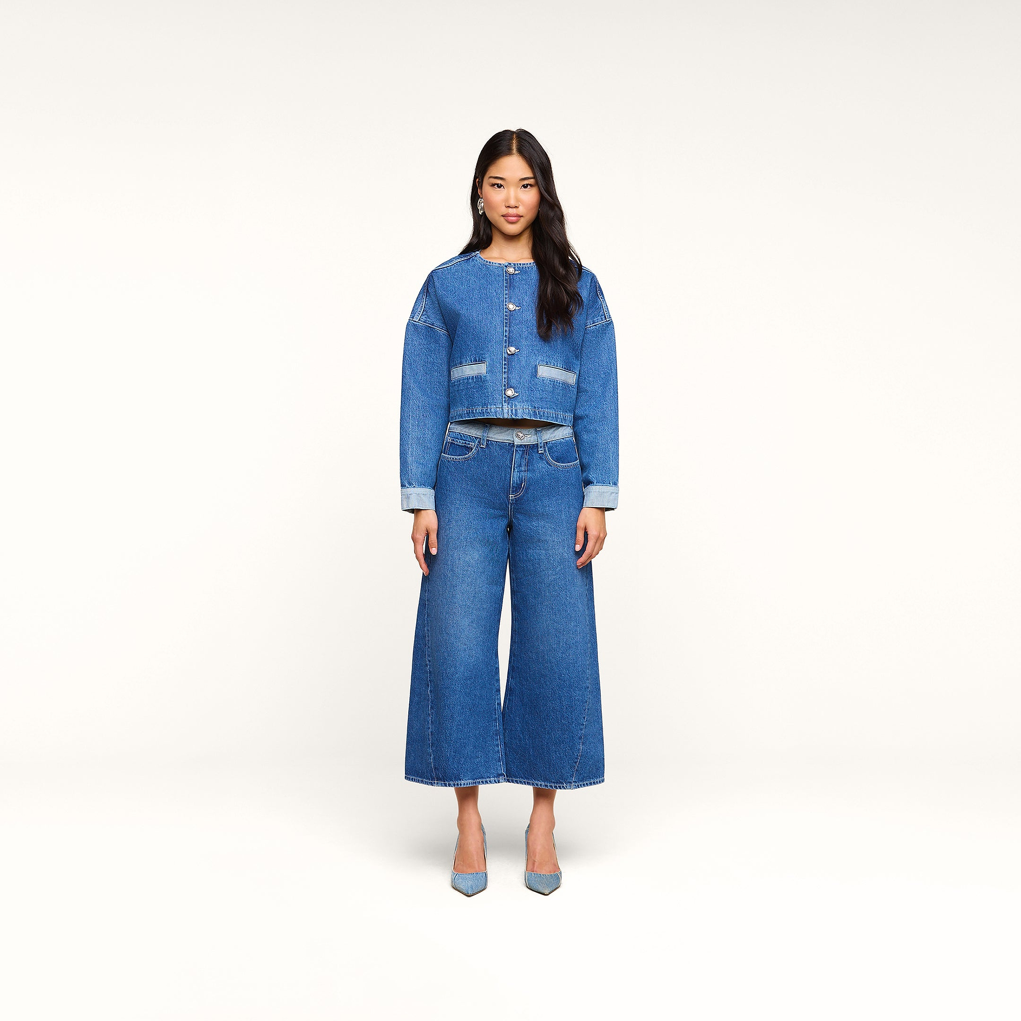 Una donna è in piedi, rivolta verso lobiettivo, indossa la giacca Verona Jean e dei jeans a gamba larga, con tacchi azzurri, su uno sfondo beige a tinta unita.