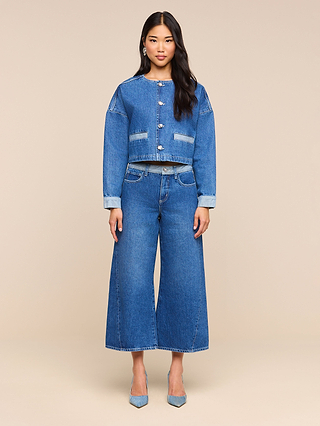 A woman stands facing forward, wearing the Verona Jean Jacket and wide-leg jeans with light blue heels against a plain beige background.