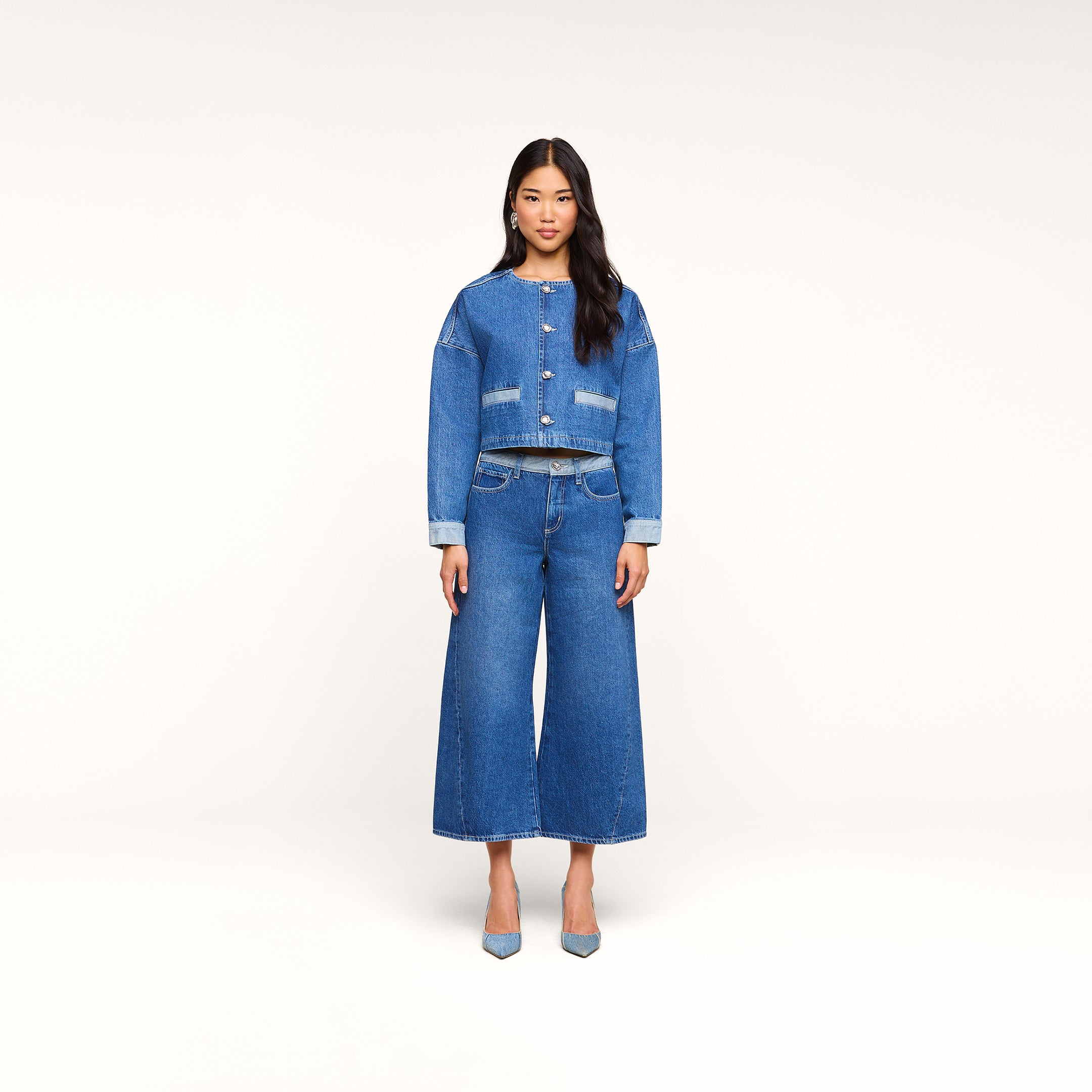 Una donna è in piedi, rivolta verso lobiettivo, indossa la giacca Verona Jean e dei jeans a gamba larga, con tacchi azzurri, su uno sfondo beige a tinta unita.