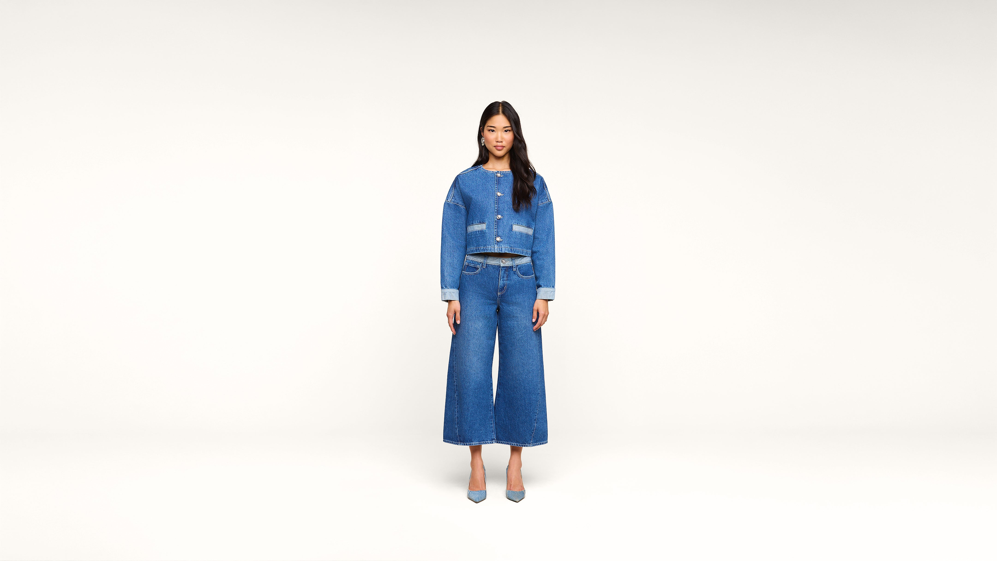 A woman stands facing forward against a plain white background, wearing a blue denim jacket, wide-leg denim pants, and matching blue heels.