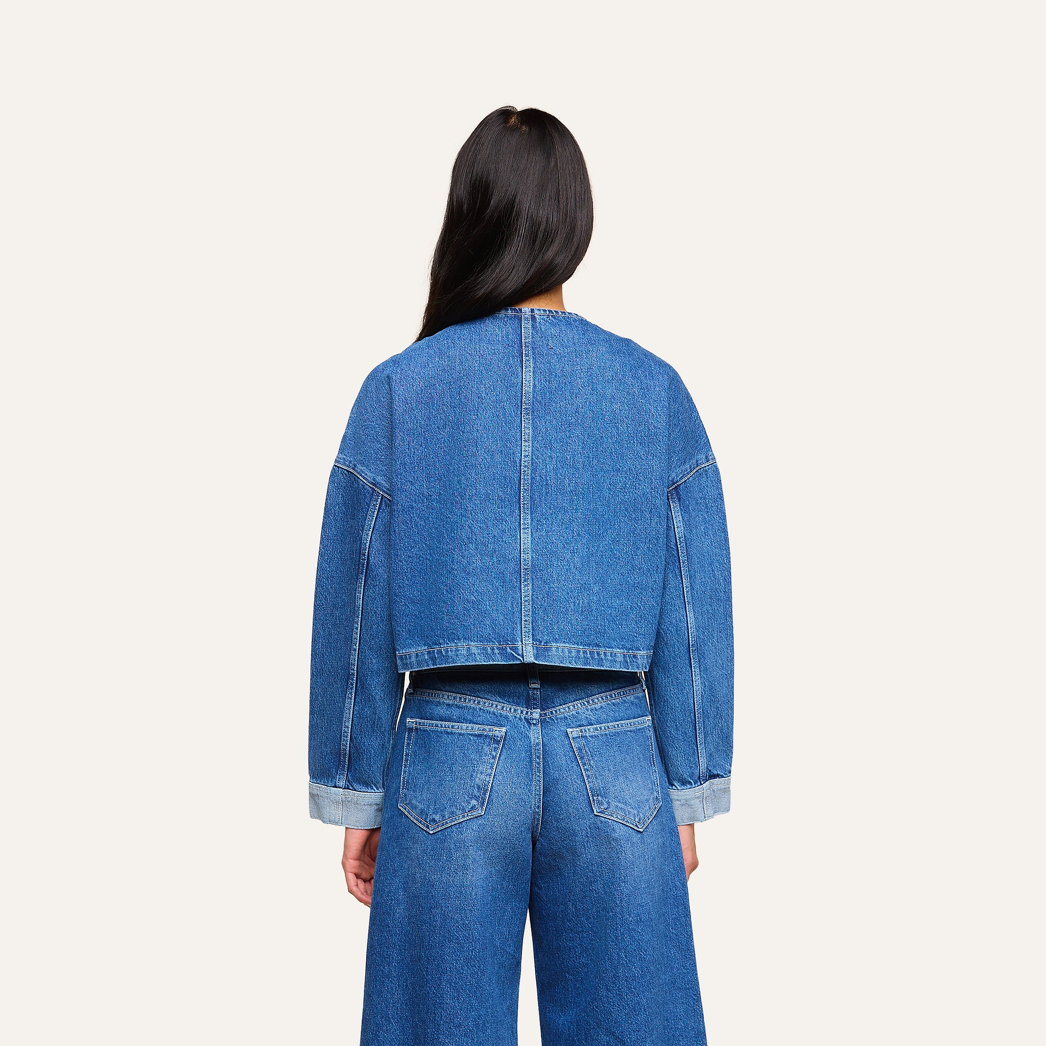 Una persona dai lunghi capelli scuri, vista di spalle, indossa la giacca di jeans Verona dal taglio oversize e dei jeans blu a vita alta su uno sfondo beige a tinta unita.