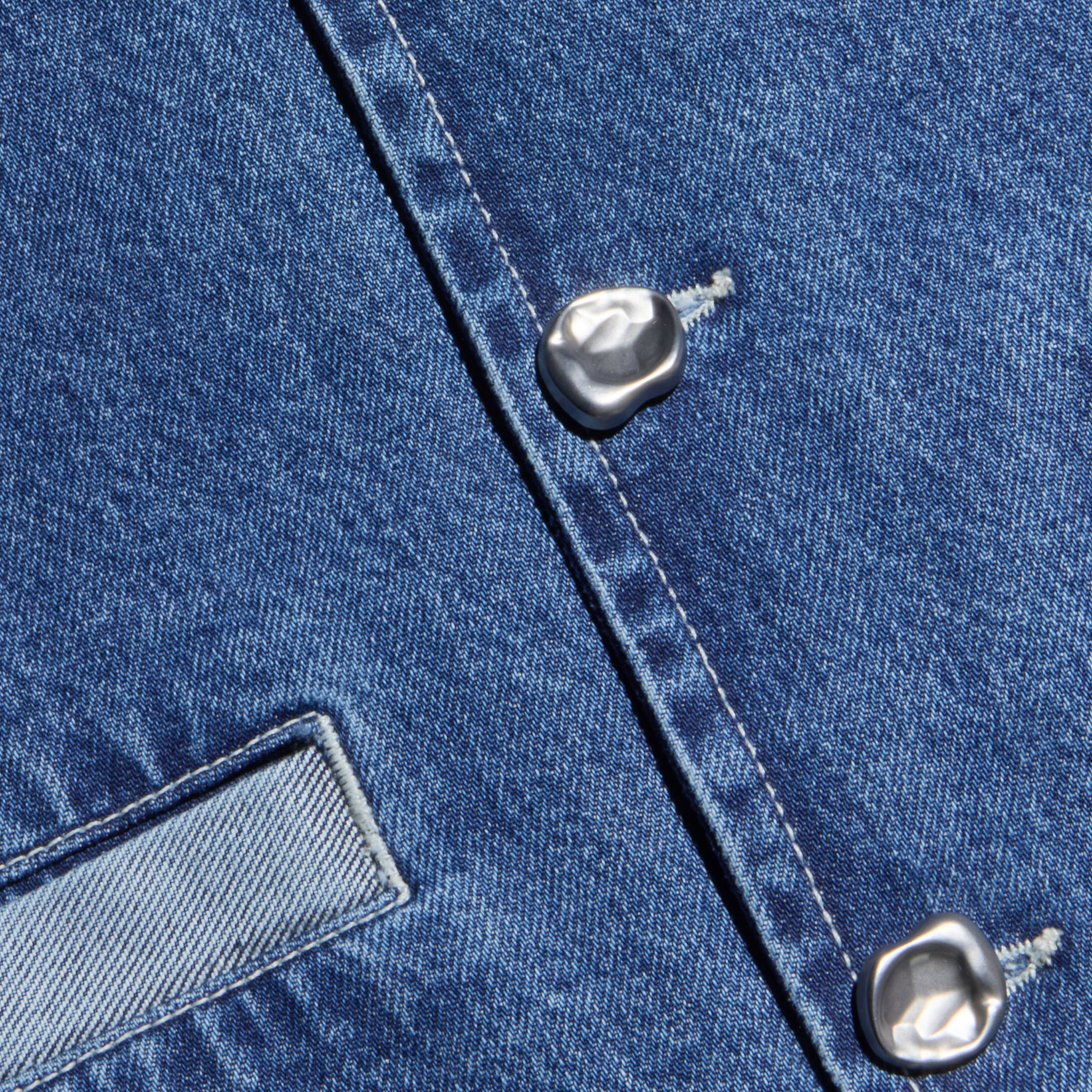 Close-up of blue denim fabric from the Verona Jean Jacket, featuring two silver buttons and a rectangular stitched pocket, highlighting the material’s texture and stitching details.