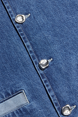 Close-up of the Verona Jean Jacket’s blue, non-stretch denim fabric, featuring silver, irregular-shaped buttons lining the seam and a small pocket partially visible on the left—details that showcase the unique structure of this collarless jacket.