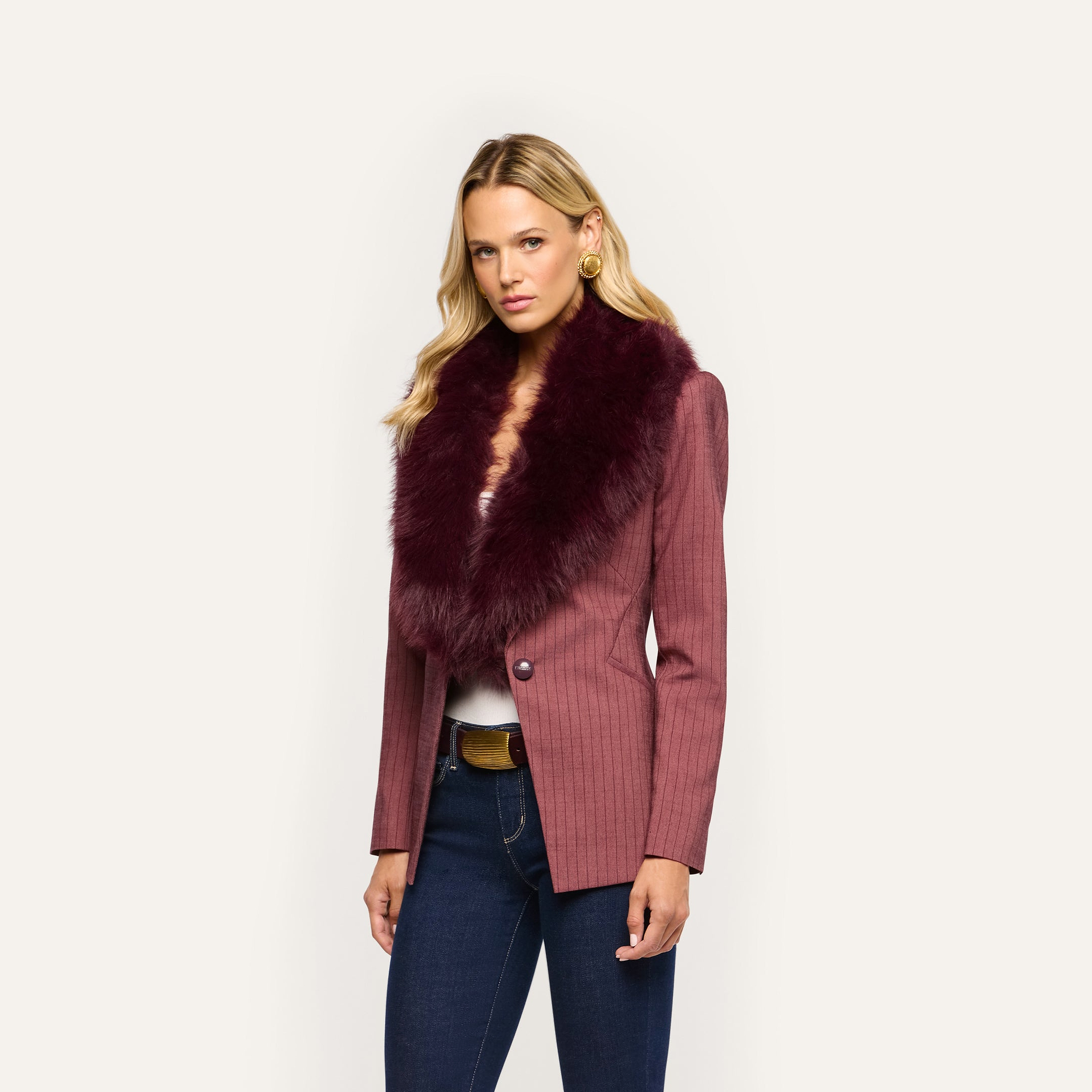 A woman wears a Sheila Blazer featuring a pinstriped pattern and a large, dark-red faux fur collar over a white top and dark blue jeans.