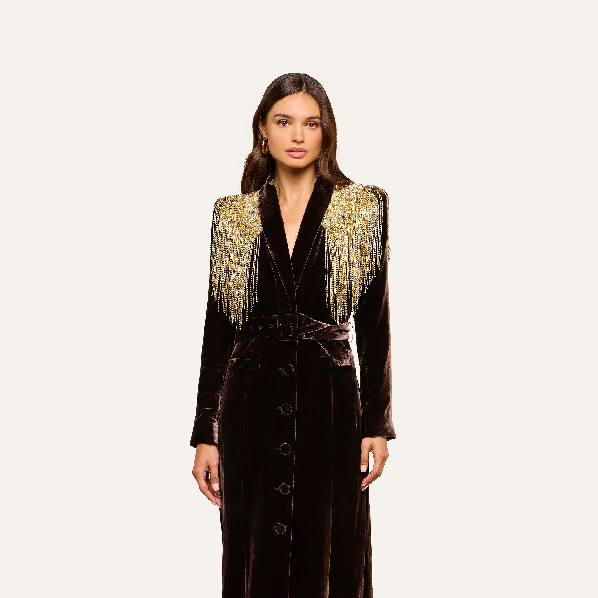 A woman stands against a plain light background, wearing the Garavani Crystal Cape—a long, dark velvet coat featuring gold beaded fringe on the shoulders and a satin collar.
