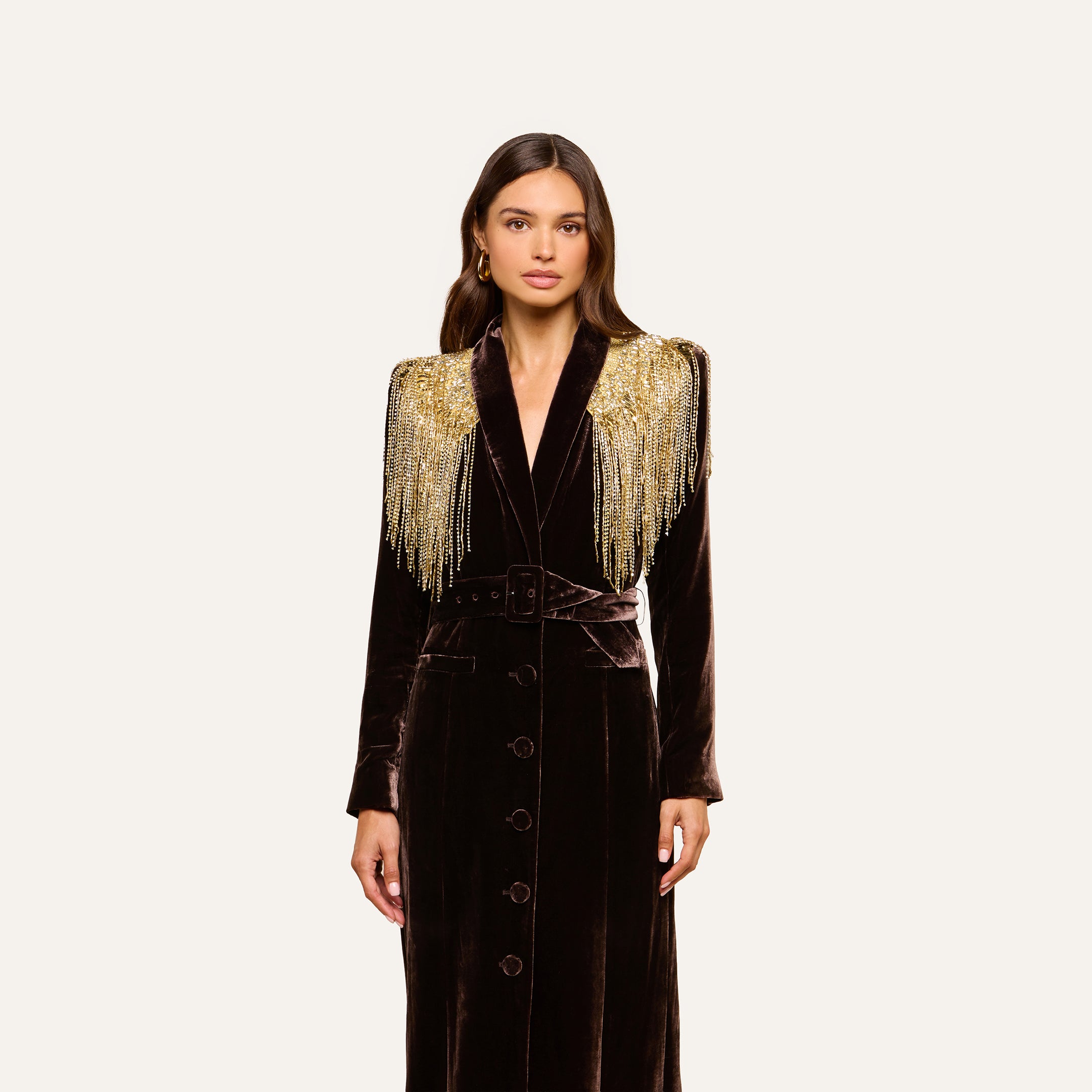 A woman stands against a plain light background, wearing the Garavani Crystal Cape—a long, dark velvet coat featuring gold beaded fringe on the shoulders and a satin collar.