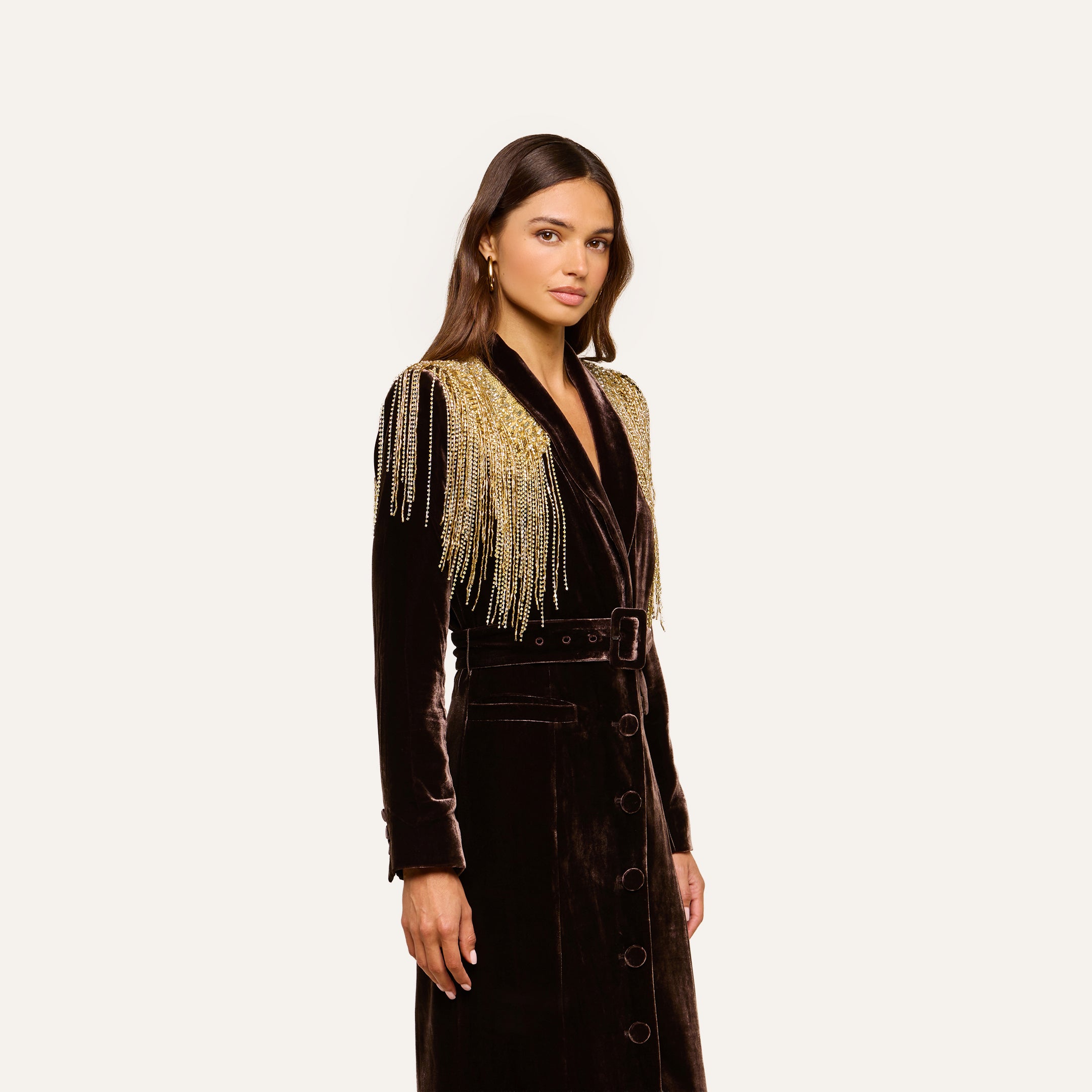A woman wearing the Garavani Crystal Cape—an elegant long black velvet coat adorned with gold beaded fringe on the shoulders and featuring a satin collar—stands against a plain light background, gazing ahead with a neutral expression.