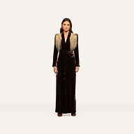 A woman stands facing forward against a plain off-white background, wearing the Garavani Crystal Cape—a long black velvet dress featuring gold fringe shoulder details, crystal embellishments, button accents down the front, and a fitted waist.