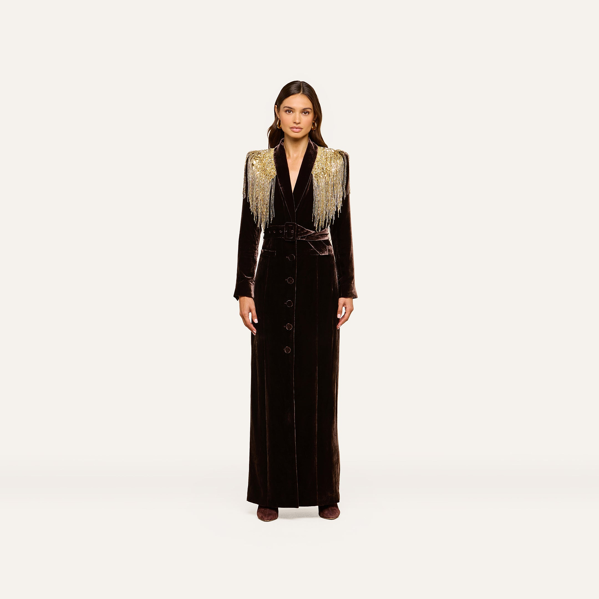 A woman stands facing forward against a plain off-white background, wearing the Garavani Crystal Cape—a long black velvet dress featuring gold fringe shoulder details, crystal embellishments, button accents down the front, and a fitted waist.