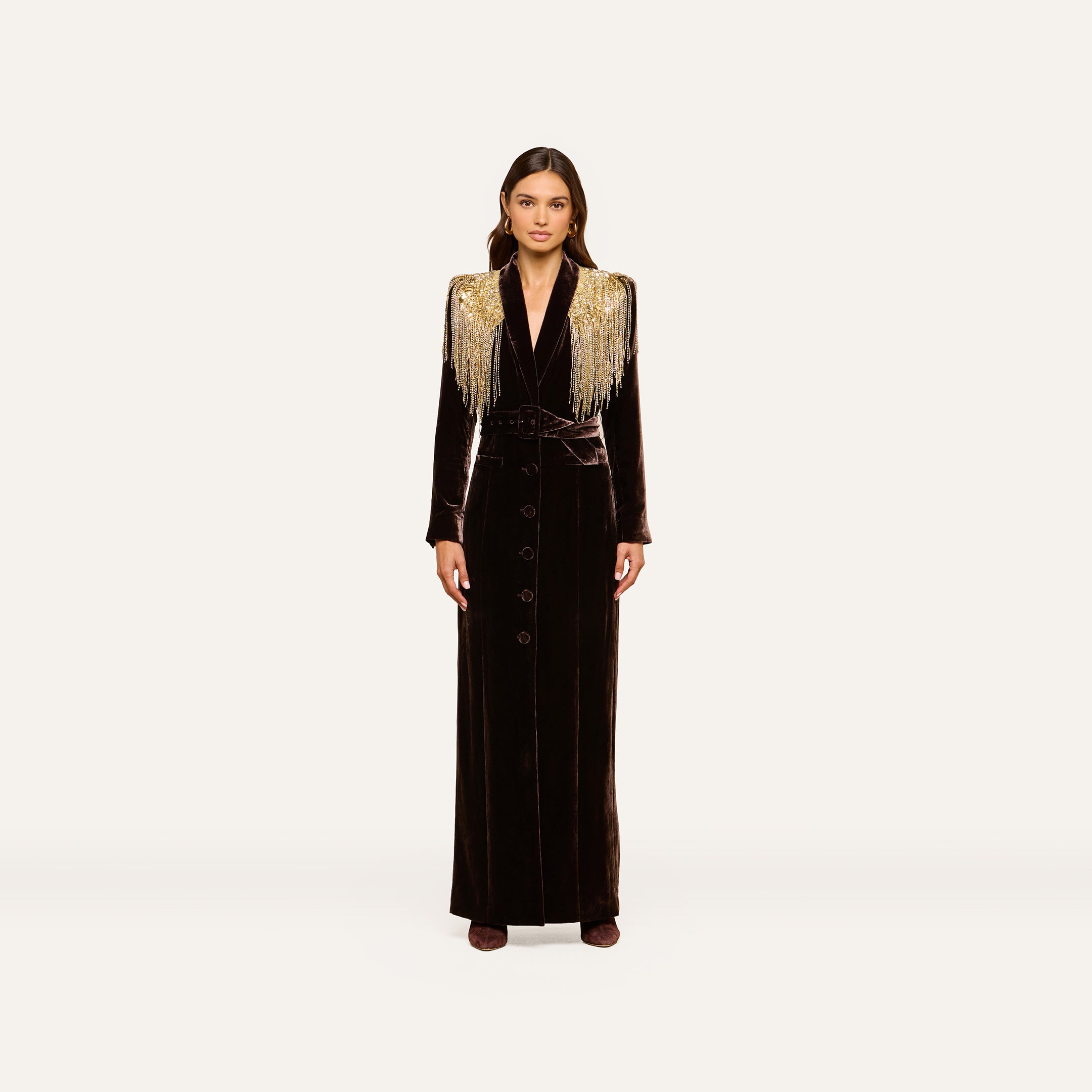 A woman stands facing forward against a plain off-white background, wearing the Garavani Crystal Cape—a long black velvet dress featuring gold fringe shoulder details, crystal embellishments, button accents down the front, and a fitted waist.