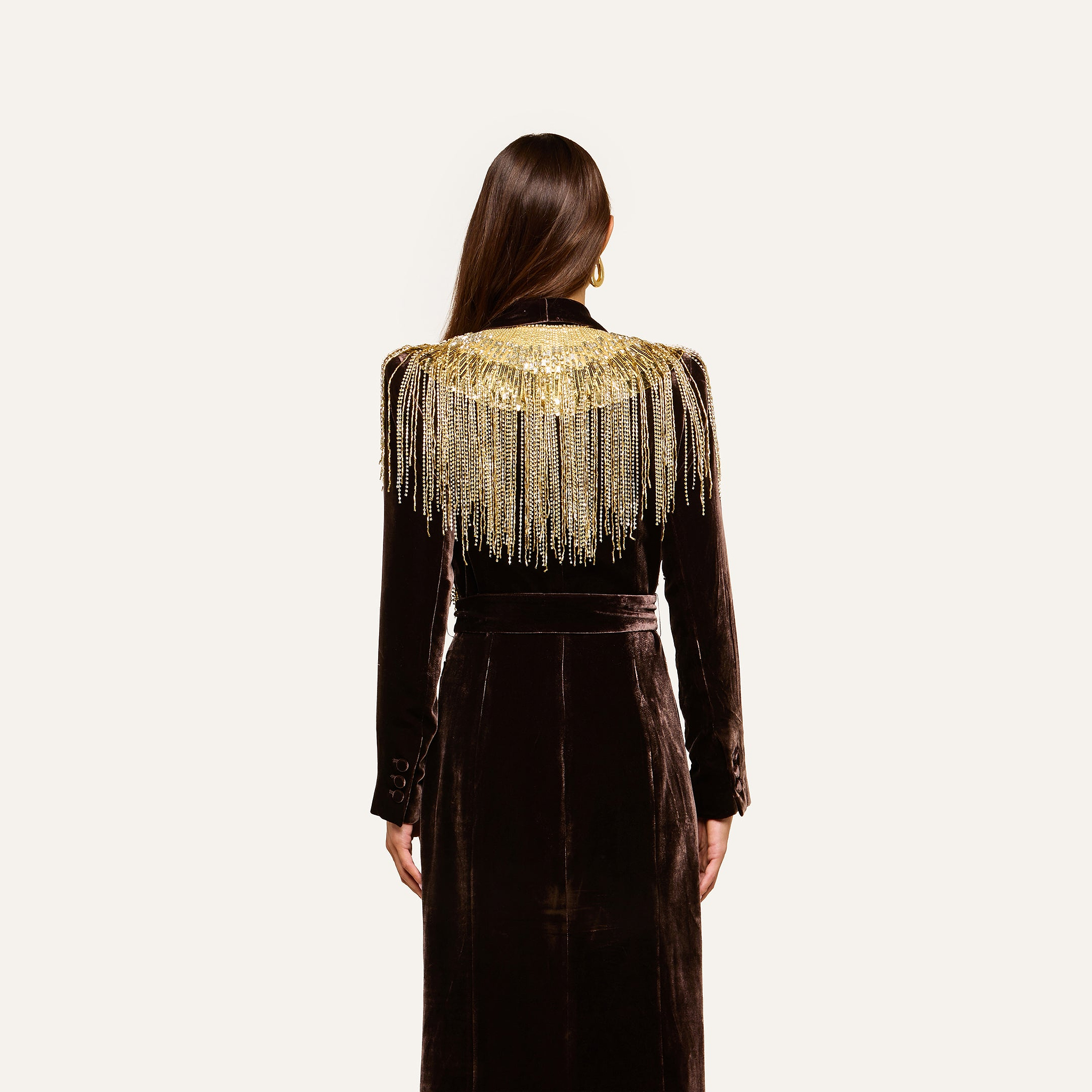 A woman with long brown hair is shown standing with her back to the camera against a plain light background, wearing the Garavani Crystal Cape—a black velvet dress featuring gold fringe detailing on the shoulders and a satin collar.