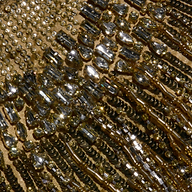 Close-up of a golden fabric, part of the Garavani Crystal Cape, adorned with shimmering sequins and beads arranged in intricate patterns that catch the light and create a sparkling, textured effect.