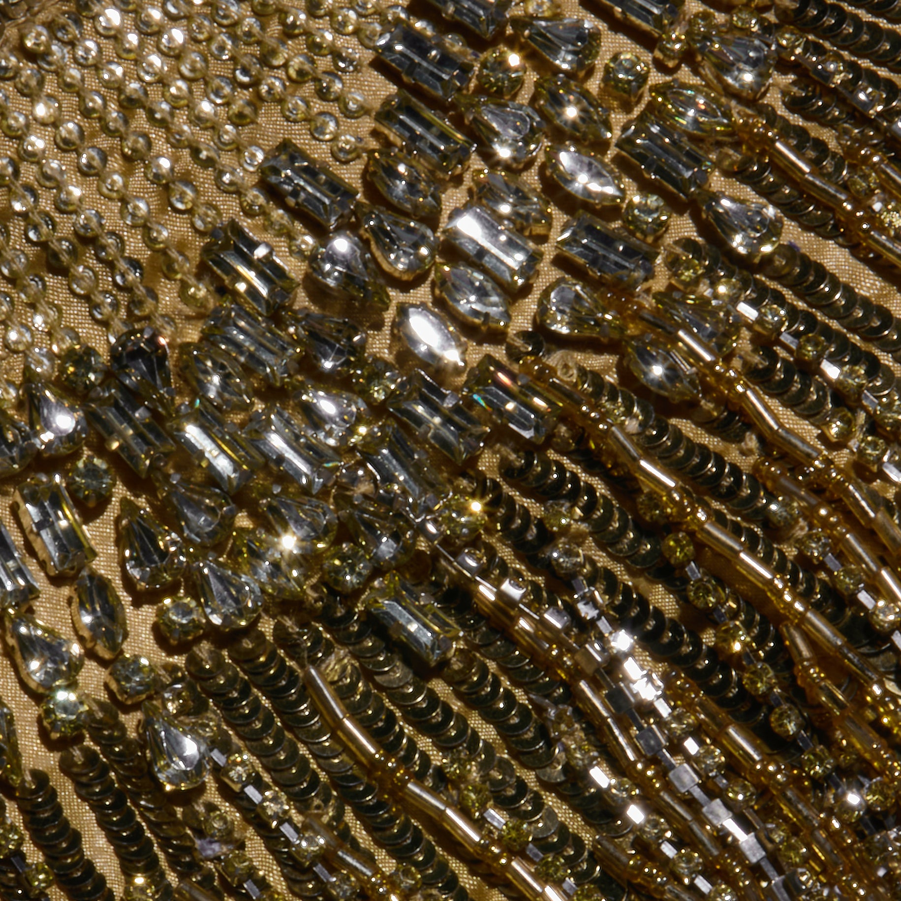 Close-up of a golden fabric, part of the Garavani Crystal Cape, adorned with shimmering sequins and beads arranged in intricate patterns that catch the light and create a sparkling, textured effect.