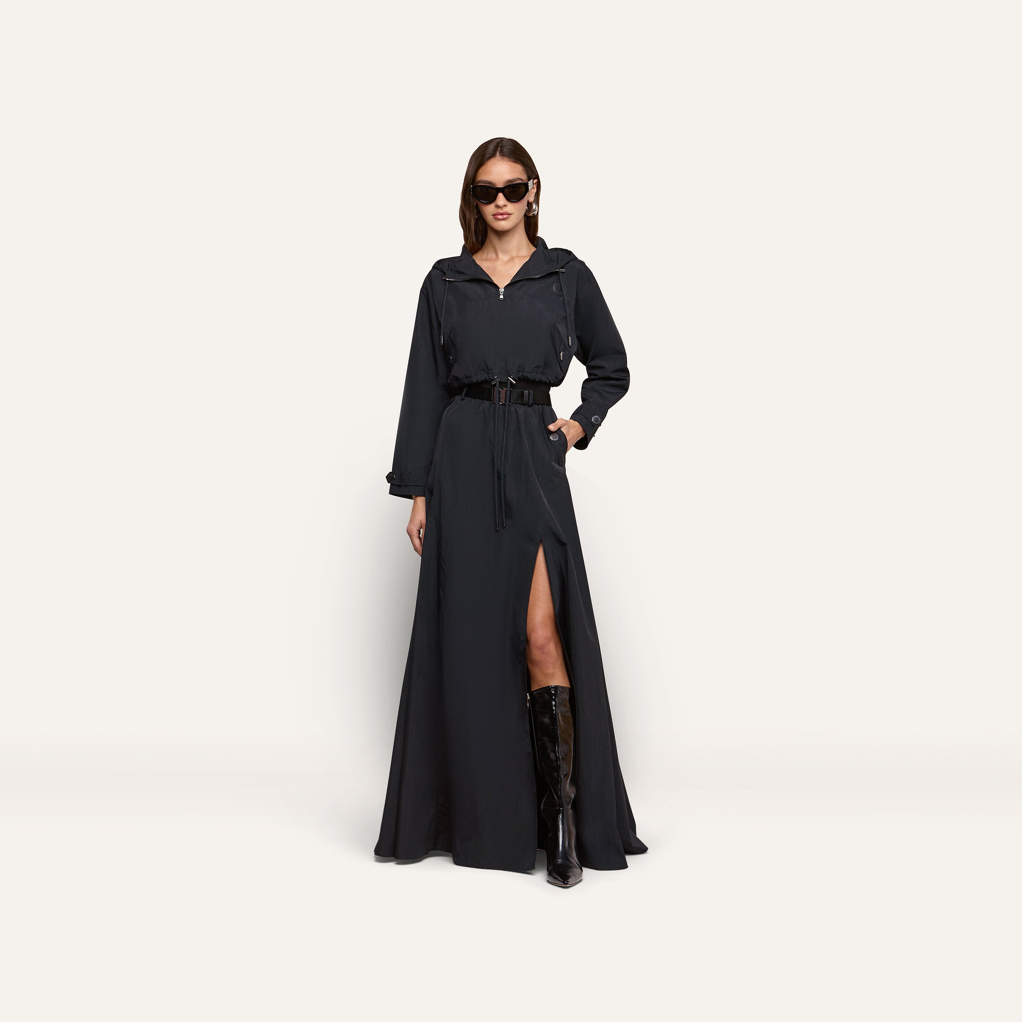 A person wearing a black Derby Windbreaker dress featuring a belted waist, front zipper, and thigh-high slit, paired with knee-high boots and sunglasses.