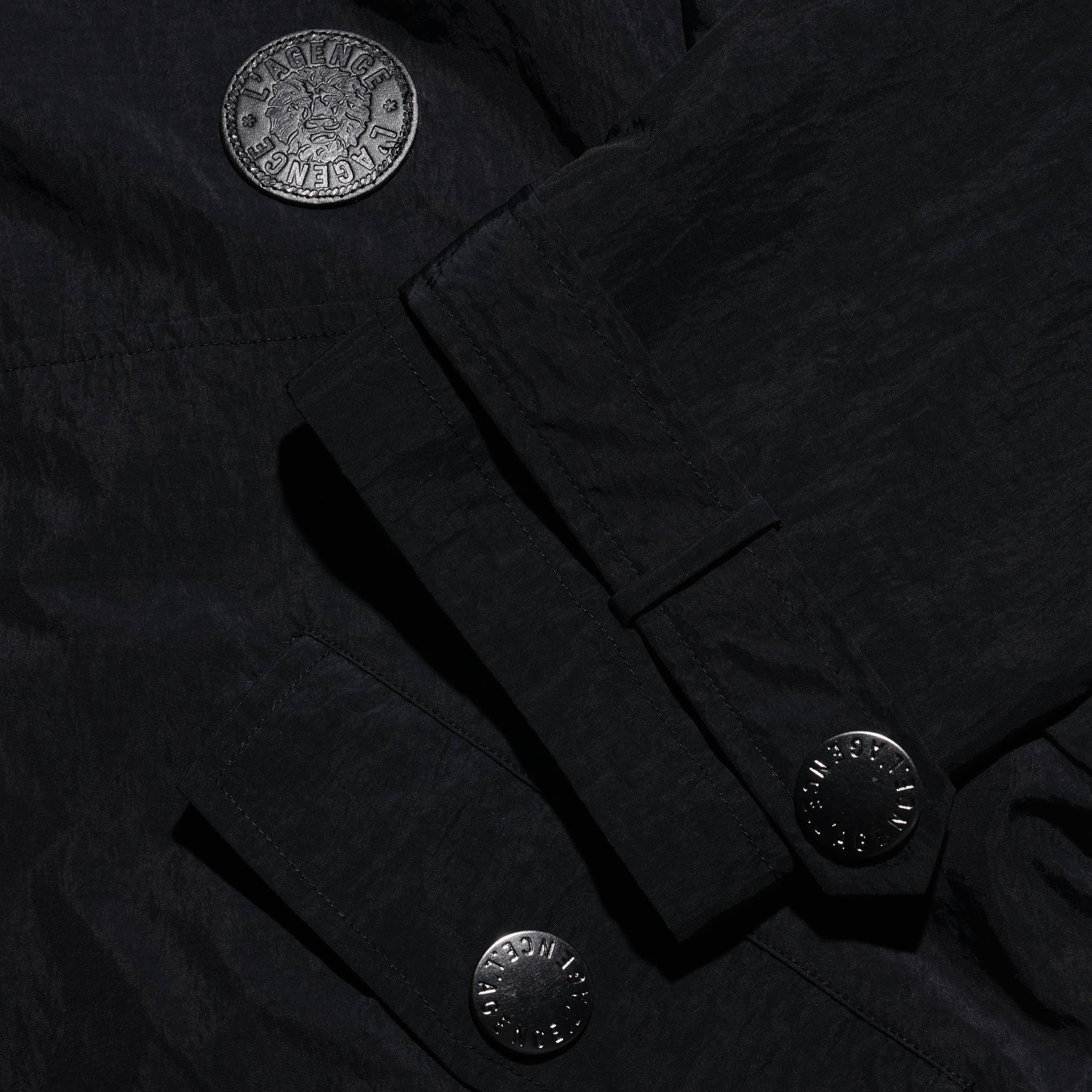 Close-up of a black, water-repellant Derby Windbreaker featuring textured fabric, a circular patch with a lion emblem, and metallic button fasteners.