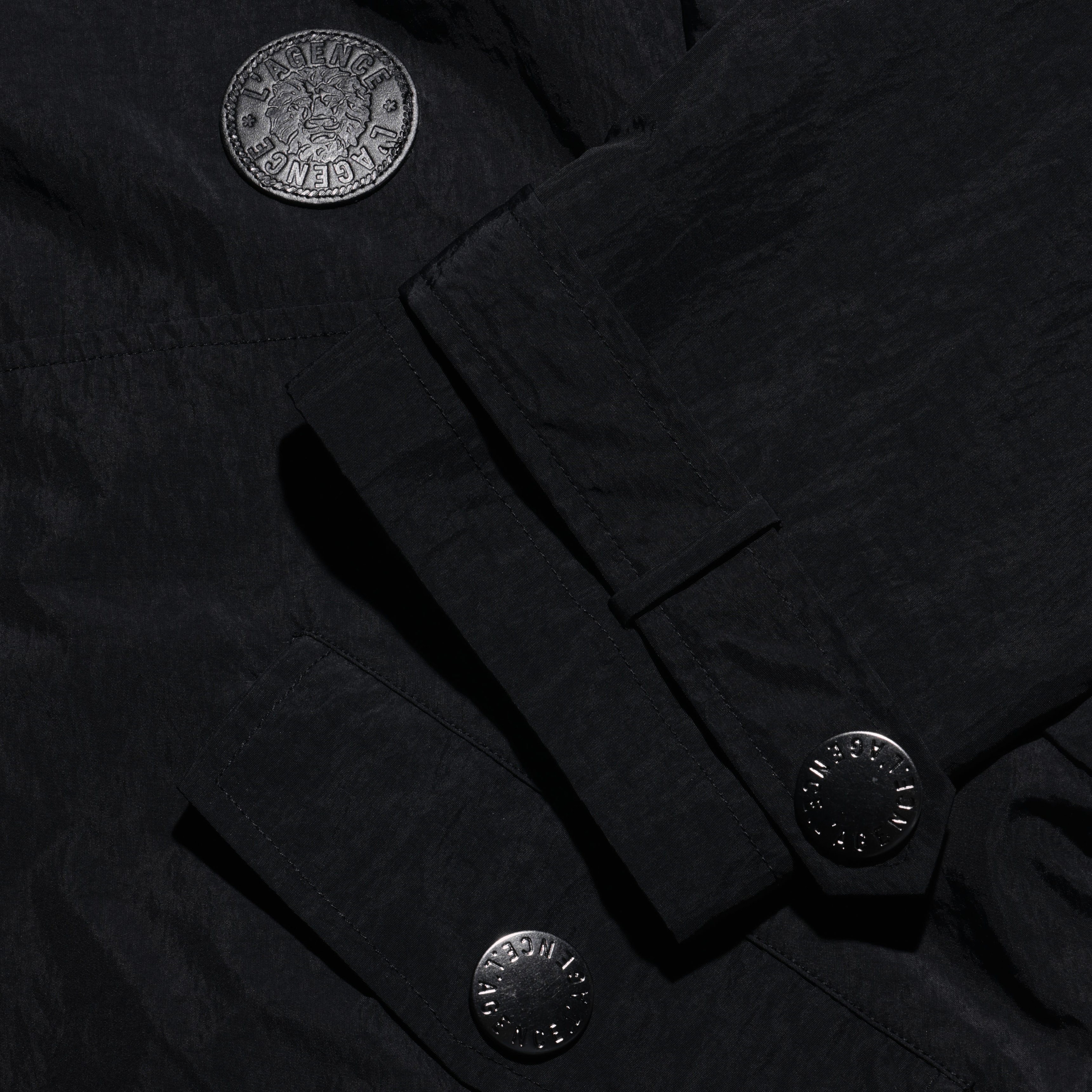 Close-up of a black, water-repellant Derby Windbreaker featuring textured fabric, a circular patch with a lion emblem, and metallic button fasteners.