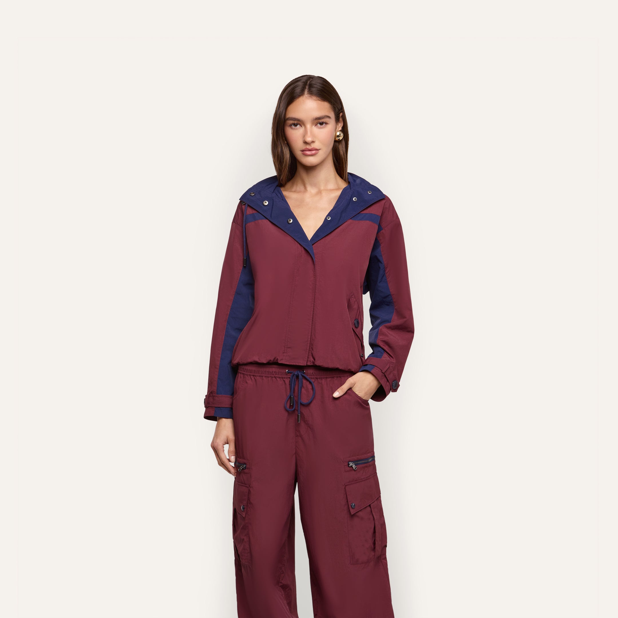 A woman wears a burgundy Geovana Windbreaker with a navy blue collar, hood, and sleeve panels, paired with matching cargo pants.